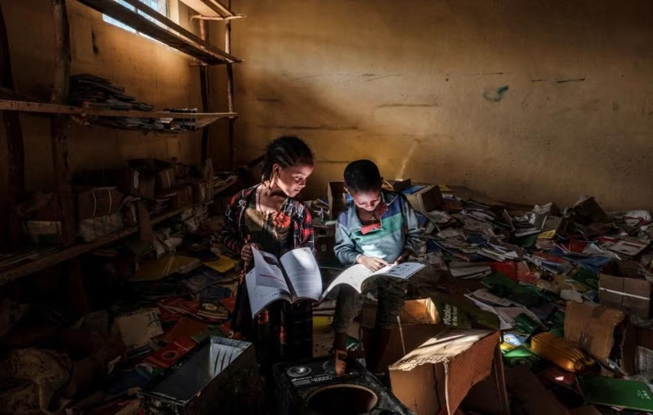 Two children reading among scattered papers in a damaged classroom